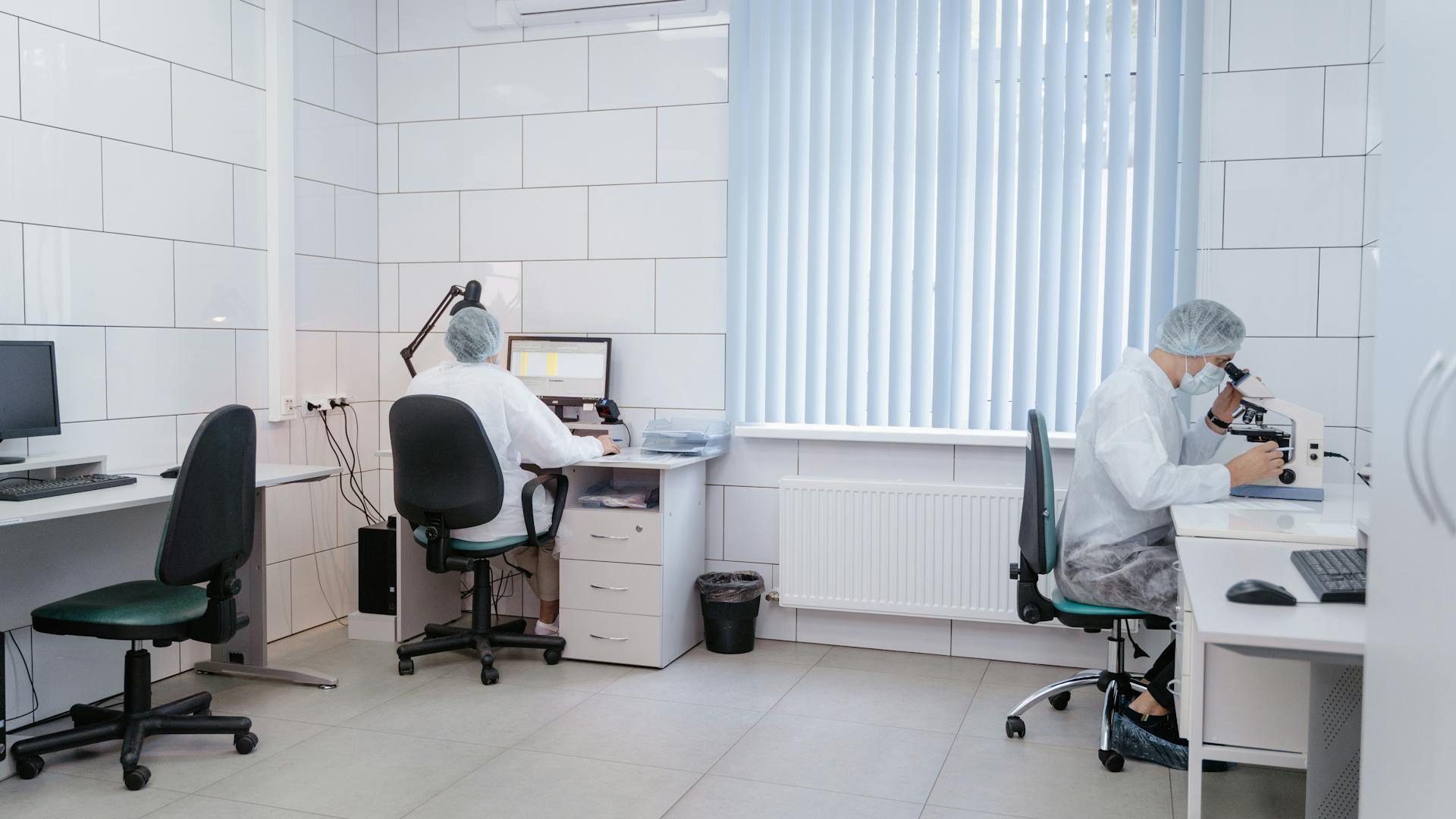 Two laboratory technicians working with computers and microscopes in a clean, modern lab.
